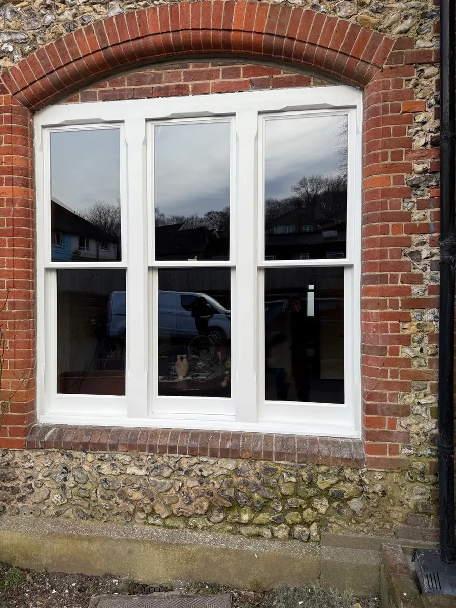 Restored white timber window under a red brick arch in a flint wall, exterior