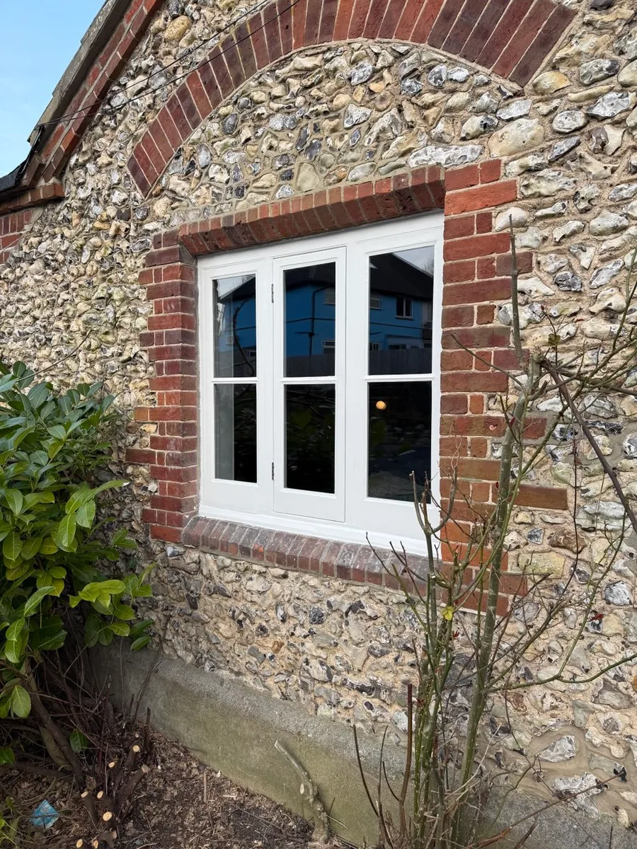 Beautifully restored white timber casement window set in a brick-arched flint wall of a period cottage